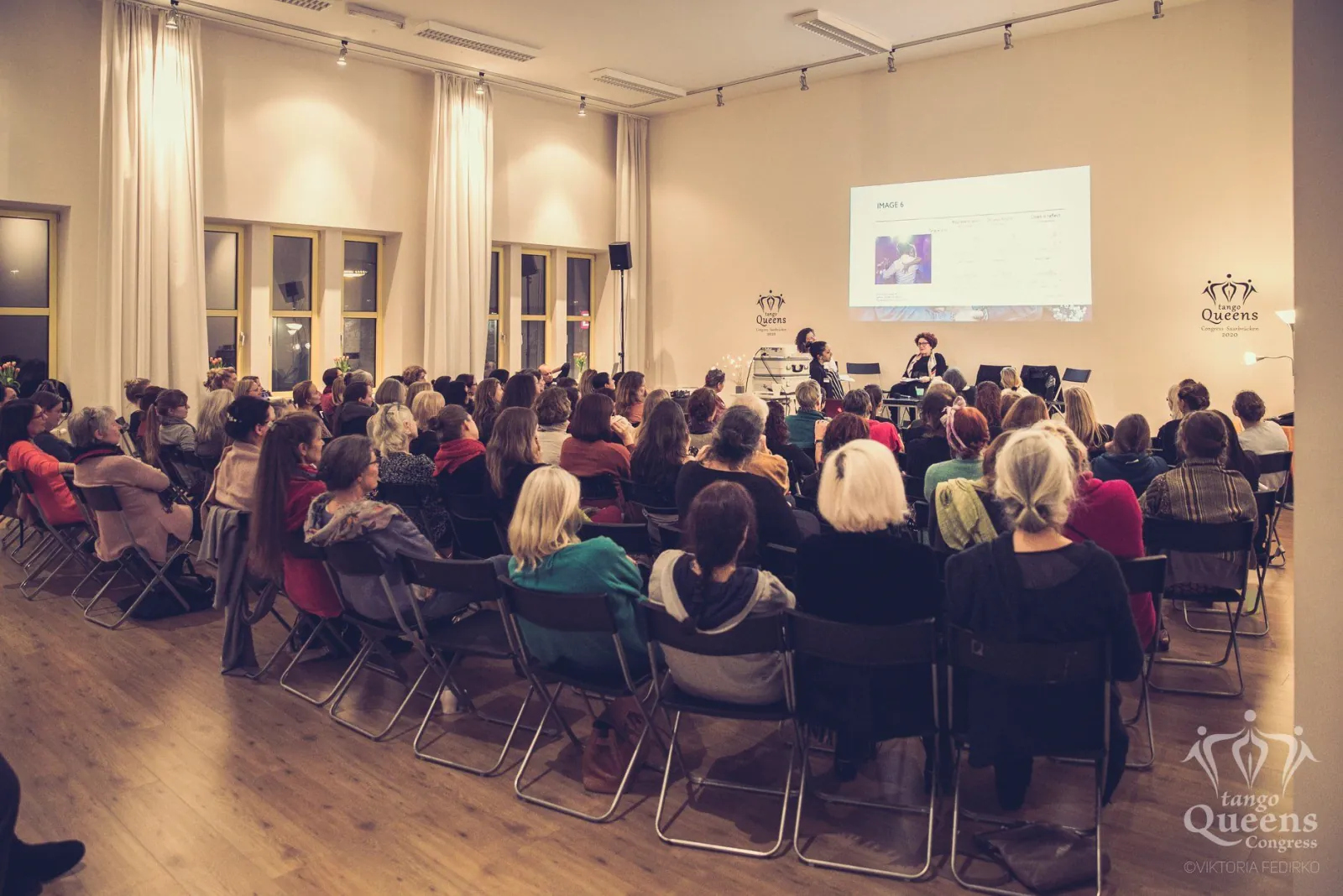 A full audience seated on chairs at a tango congress, facing two speakers beside a projection screen.