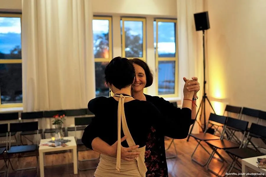 Two dancers in a close embrace by tall windows at dusk, warm light and candlelit tables behind them.