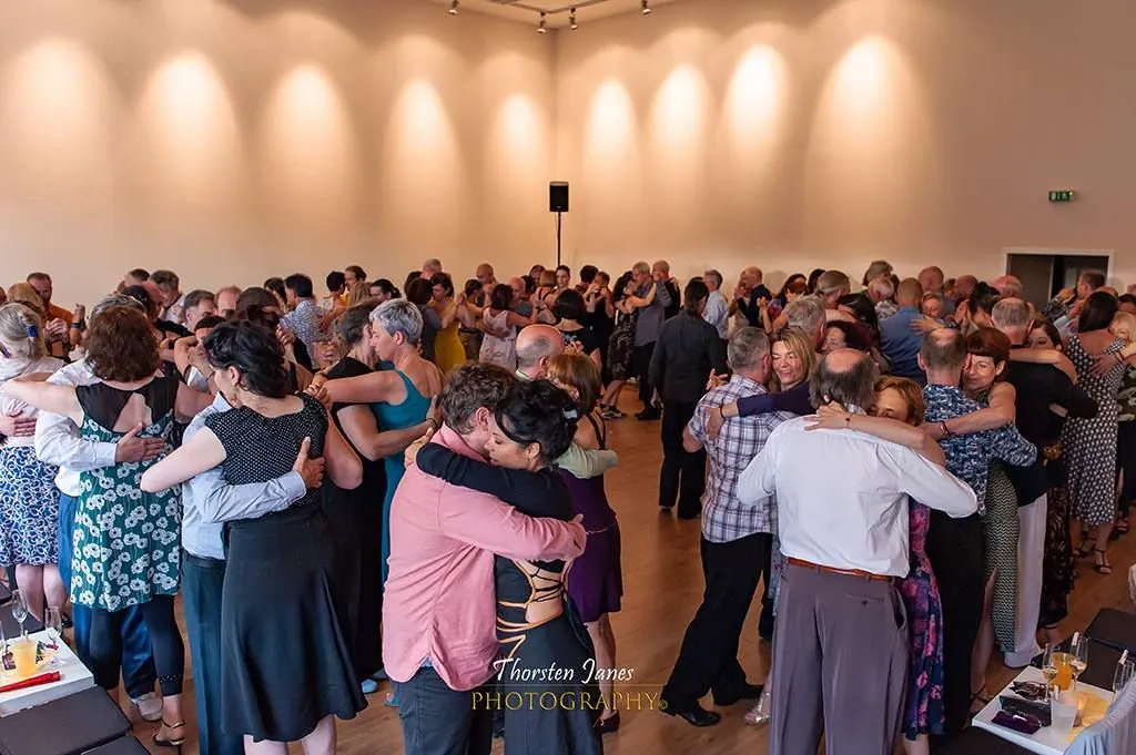 A crowded milonga with many couples dancing cheek to cheek across the floor.