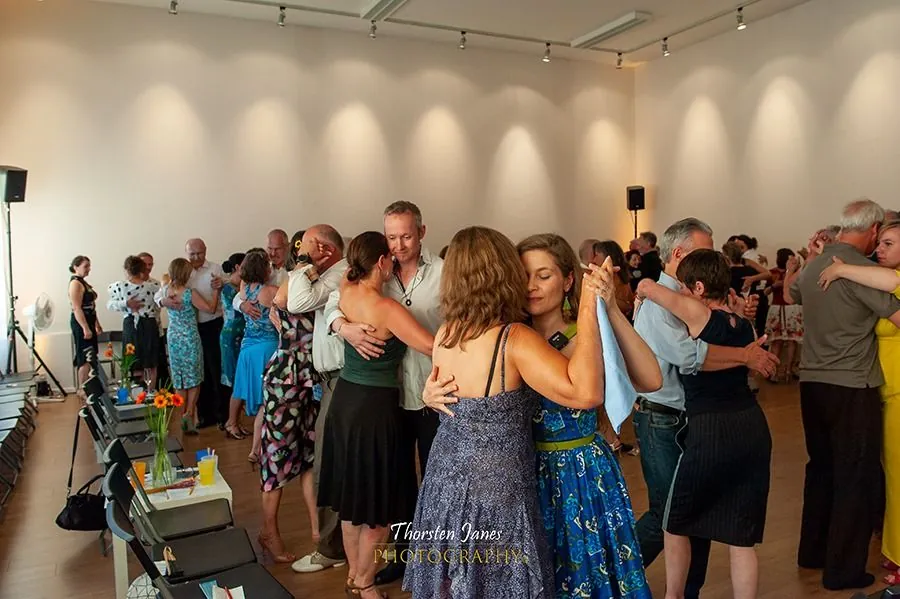 A well-attended evening milonga, many couples dancing cheek to cheek, side chairs with flowers and glasses.