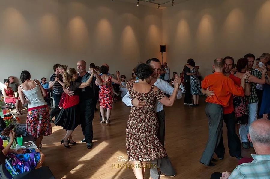 A lively milonga seen across the floor, many couples dancing while others watch from the edges.