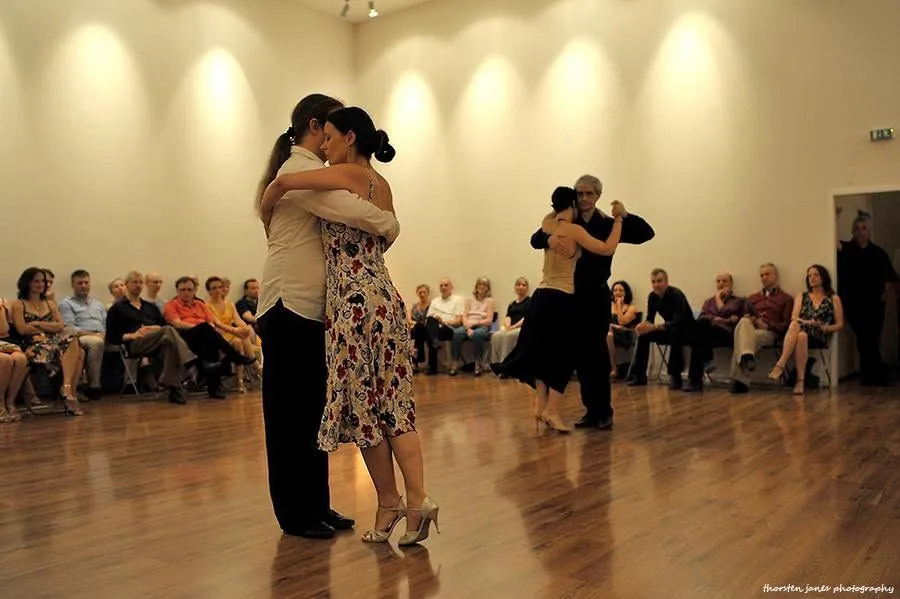A couple dancing tango in a close embrace on an open floor while seated guests watch from the sides.