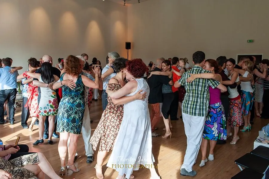 A busy summer milonga, many couples in bright dresses dancing together on the floor.