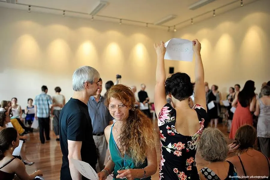 People mingling at a warmly lit social event, one person holding a handwritten sign above the crowd.