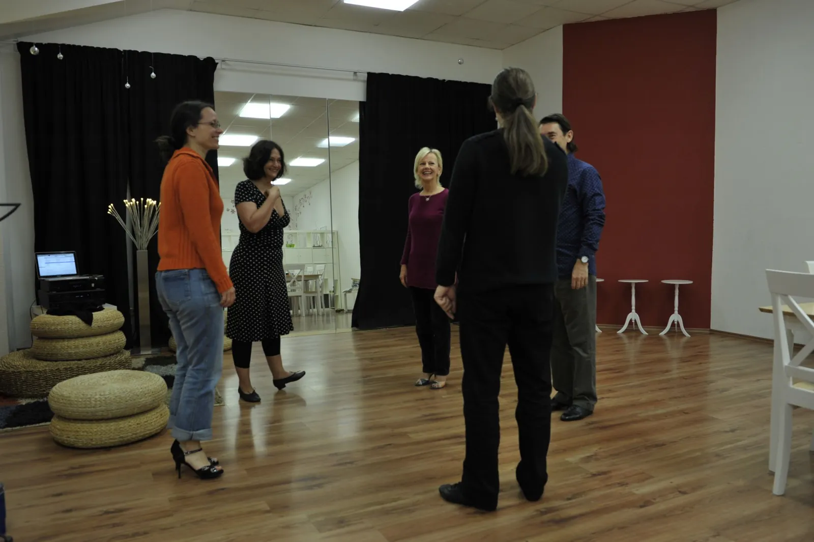 A handful of dancers laughing together during a class in the studio, mirror and red wall behind them.