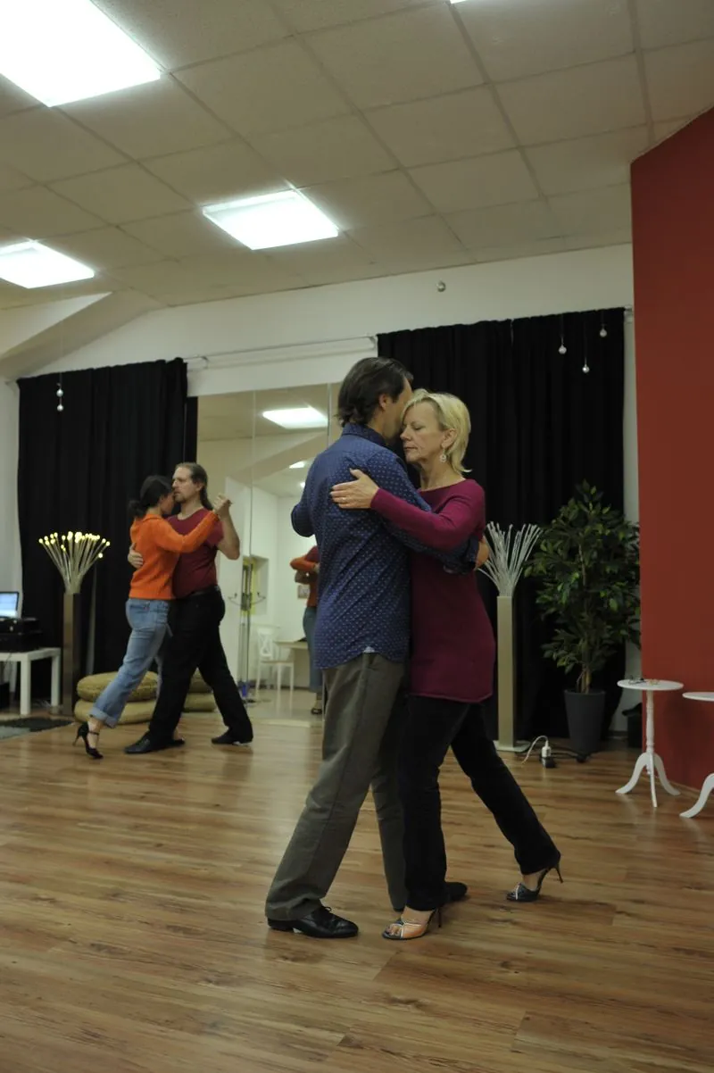 A couple dancing tango in the studio by the red wall, a second couple reflected in the mirror behind them.