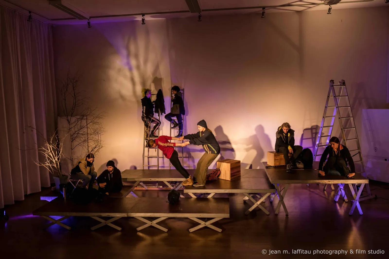 A theatre ensemble on low staging under purple light, some climbing ladders, others reaching to help a performer up.