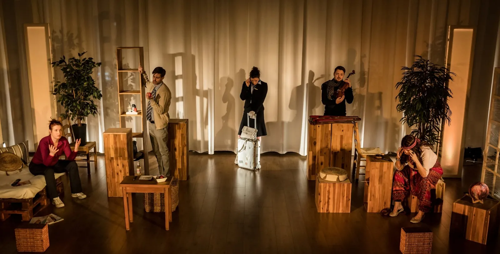 Five performers in a warmly lit theatre scene, spread across a sparse set with plants, a wooden shelf and a suitcase.