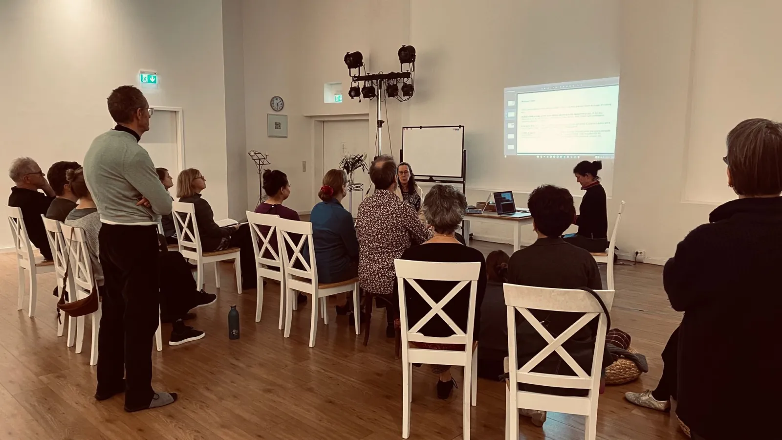 A small group on white chairs following a workshop in the studio, facing a presenter and a projected screen.
